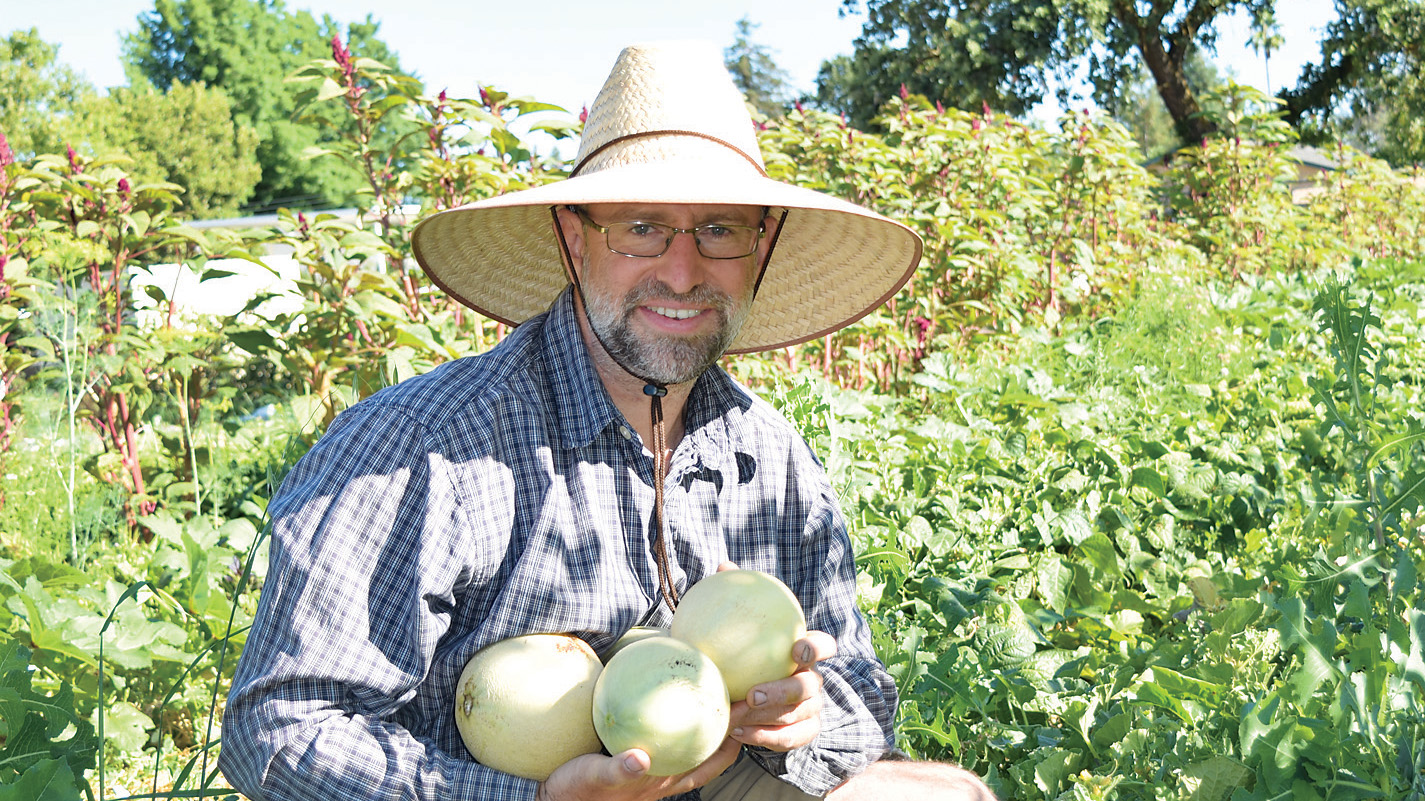 Randy Stannard in a straw hat holding three melons in the Root 64 field, flowering amaranth behind him